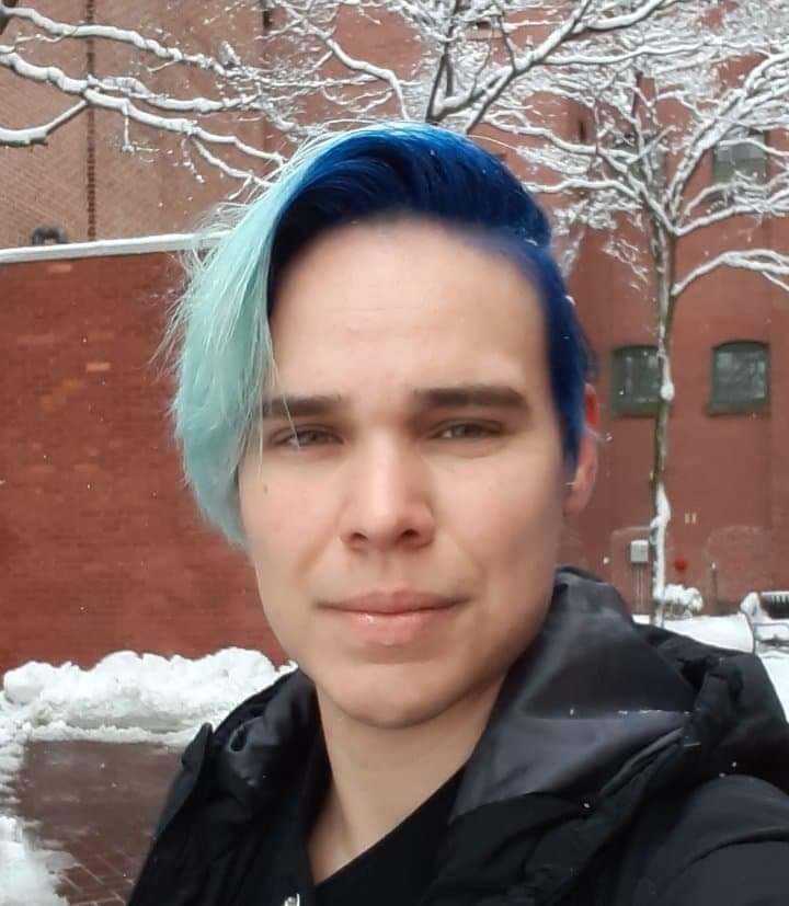 A headshot of a person with blue hair standing in front of brick building. He is looking at the camera. Snow is falling.